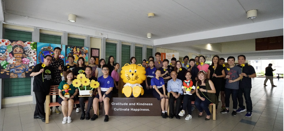 Group of people posing with a yellow lion mascot and sunflowers near a sign reading Gratitude and Kindness Cultivate Happiness.